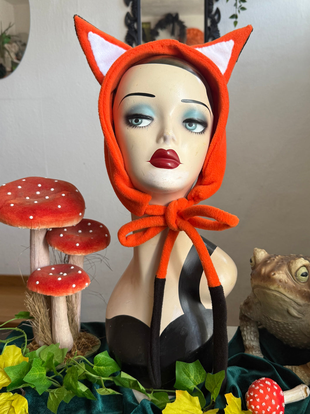 Mannequin head wearing an orange fox fleece headband with decorative mushrooms and plants in the background.
