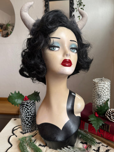 Mannequin head with black wig, red lipstick, and Krampus devil horns on a festive winter background.