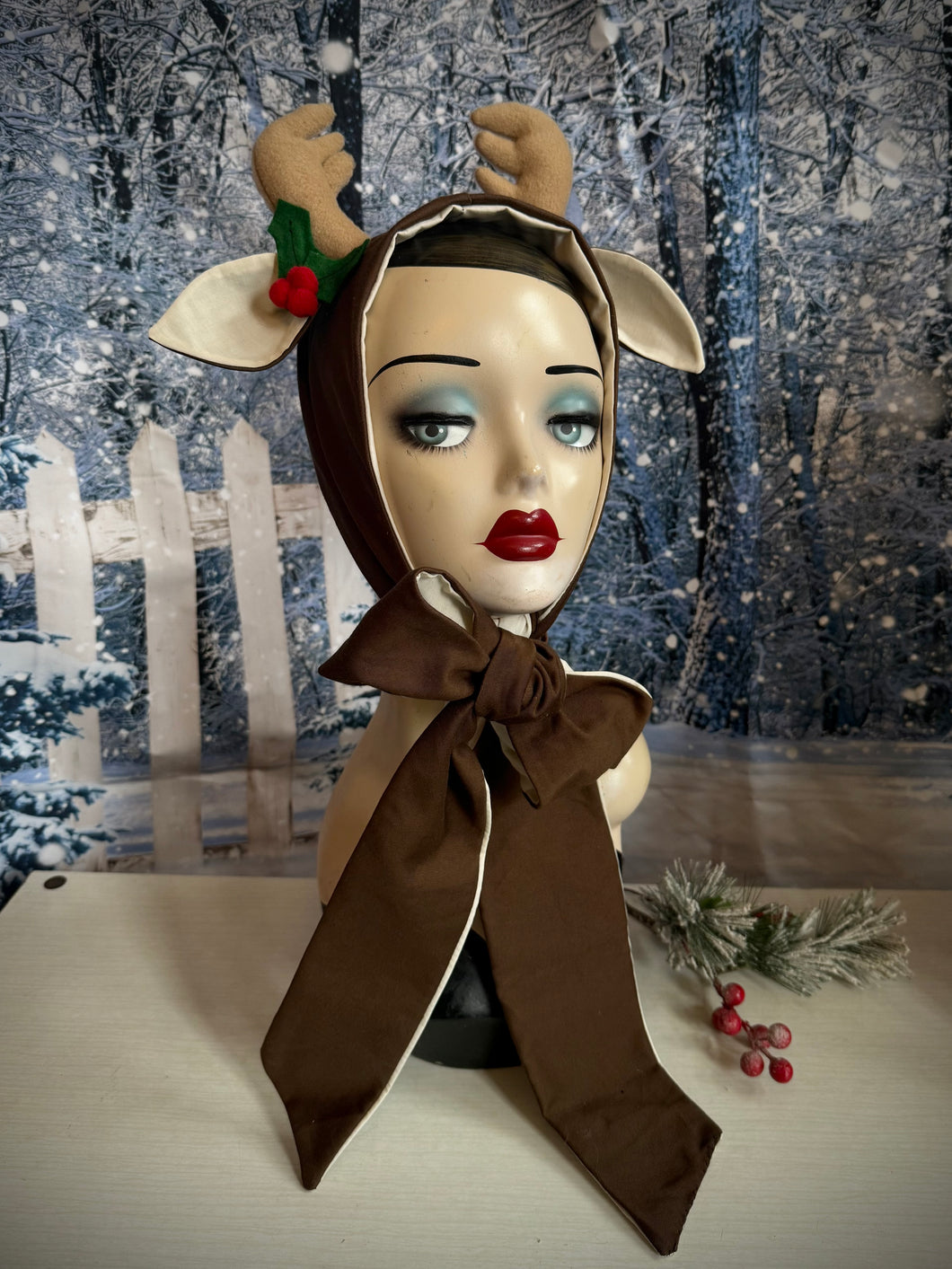 Mannequin head wearing a reindeer costume with a brown scarf against a snowy background.