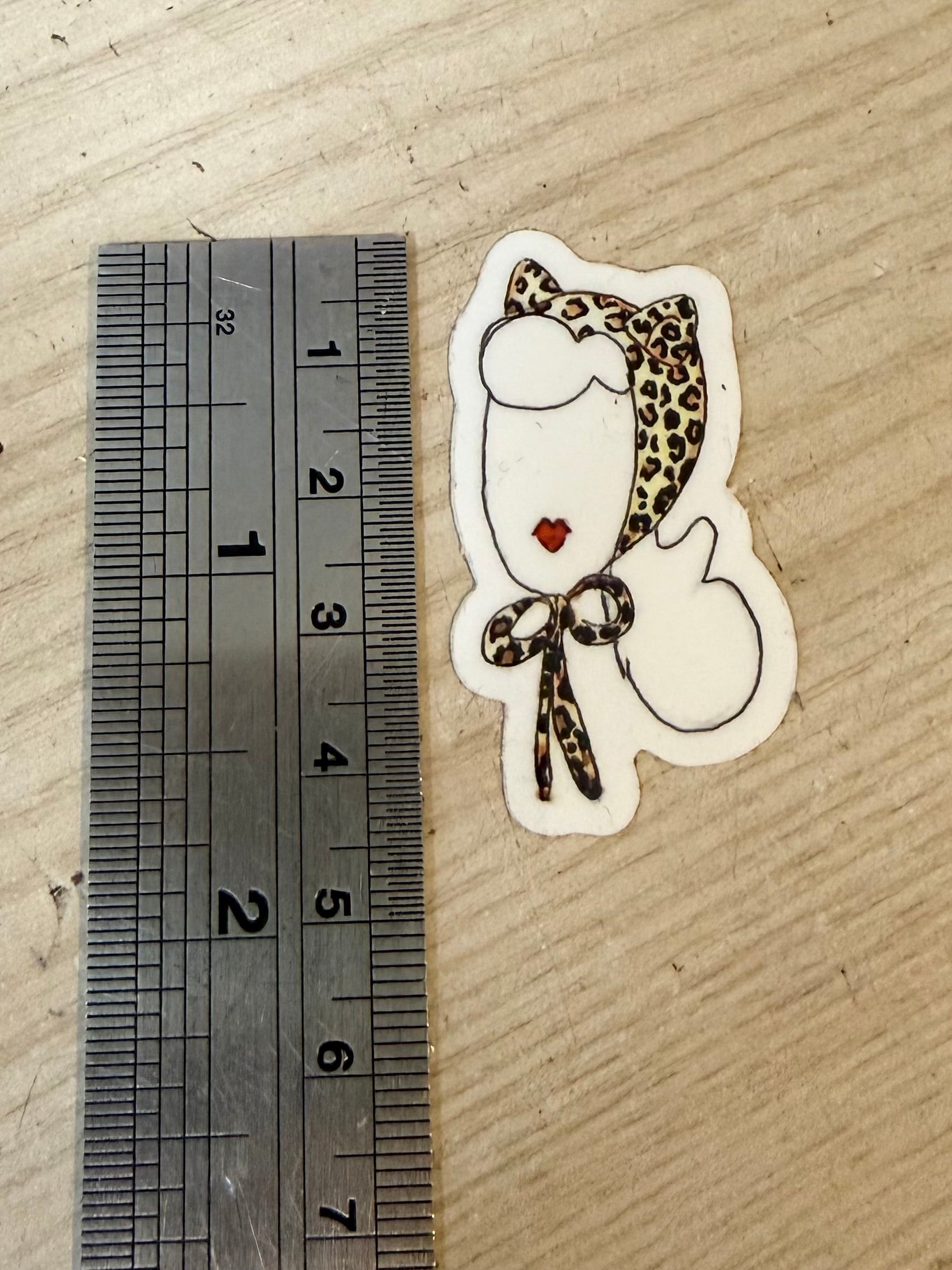 Sticker of a cat wearing a leopard print bonnet next to a ruler on cardboard