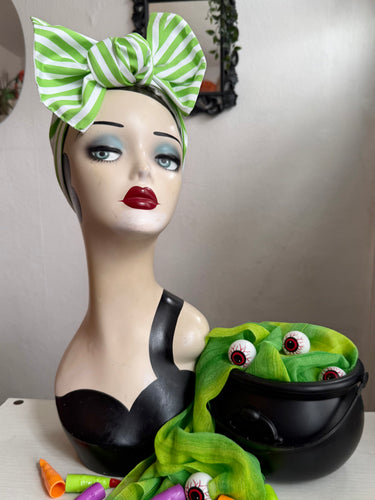 Mannequin head with green and white striped headscarf, black top, and green scarf with eyes in a black cauldron.