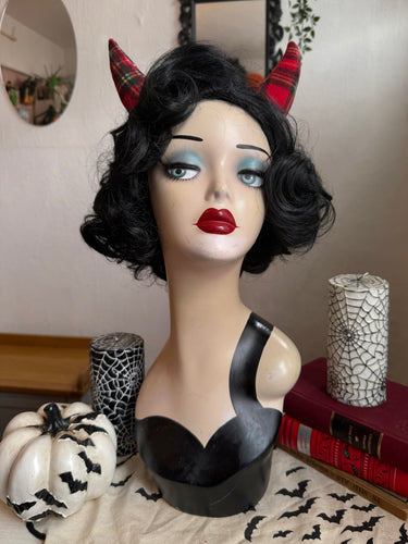 Mannequin head with black wig, red lipstick, and red devil horns against a neutral background.