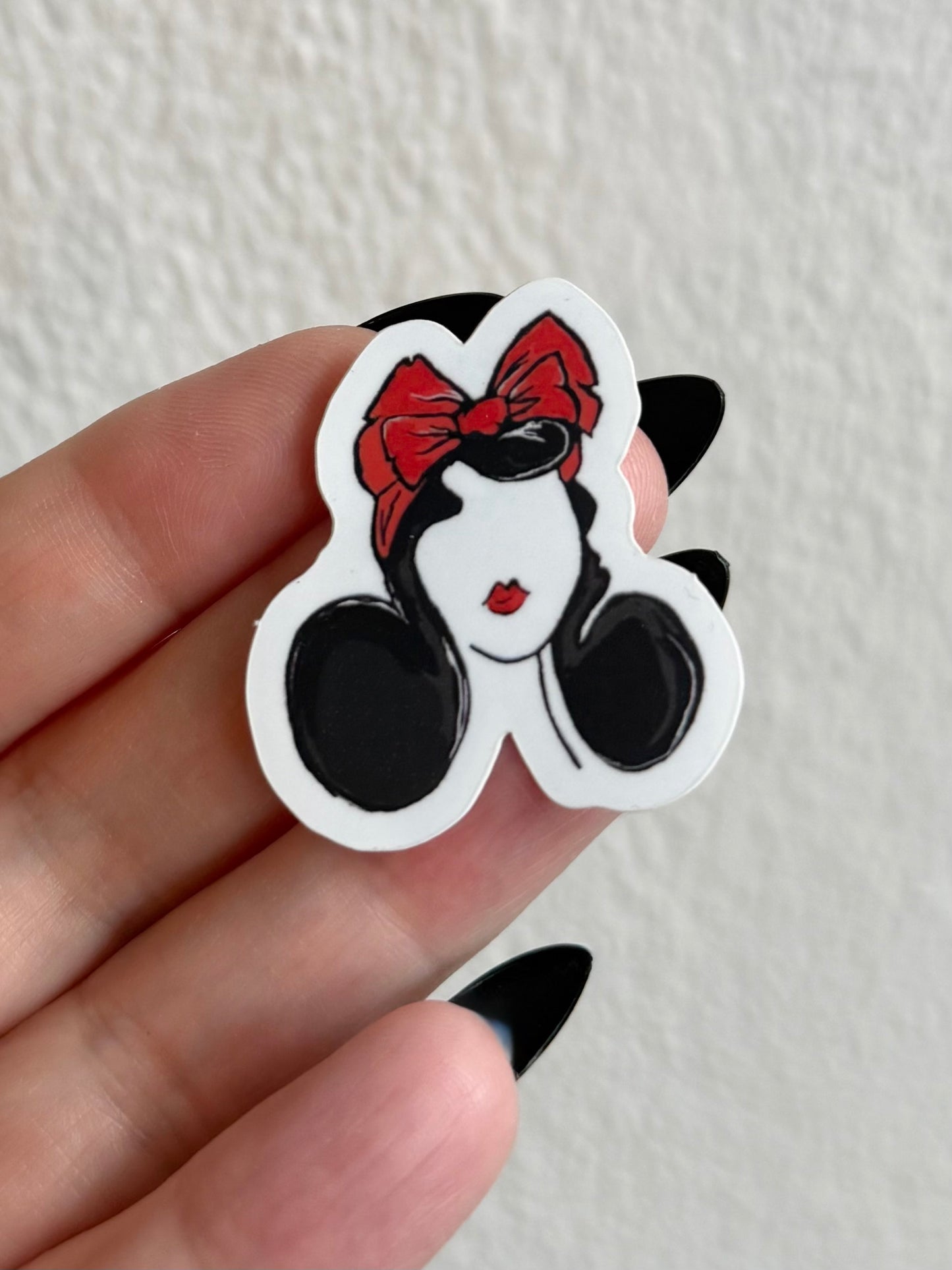 Cartoon character sticker with a red bow held between fingers against a neutral background