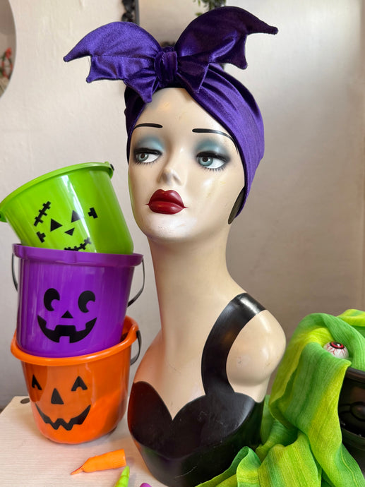 Mannequin head with purple bat headband, Halloween buckets, and green fabric on a neutral background