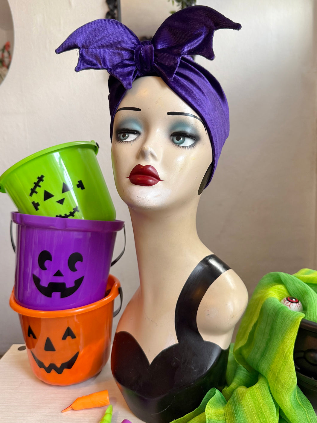 Mannequin head with purple bat headband, Halloween buckets, and green fabric on a neutral background
