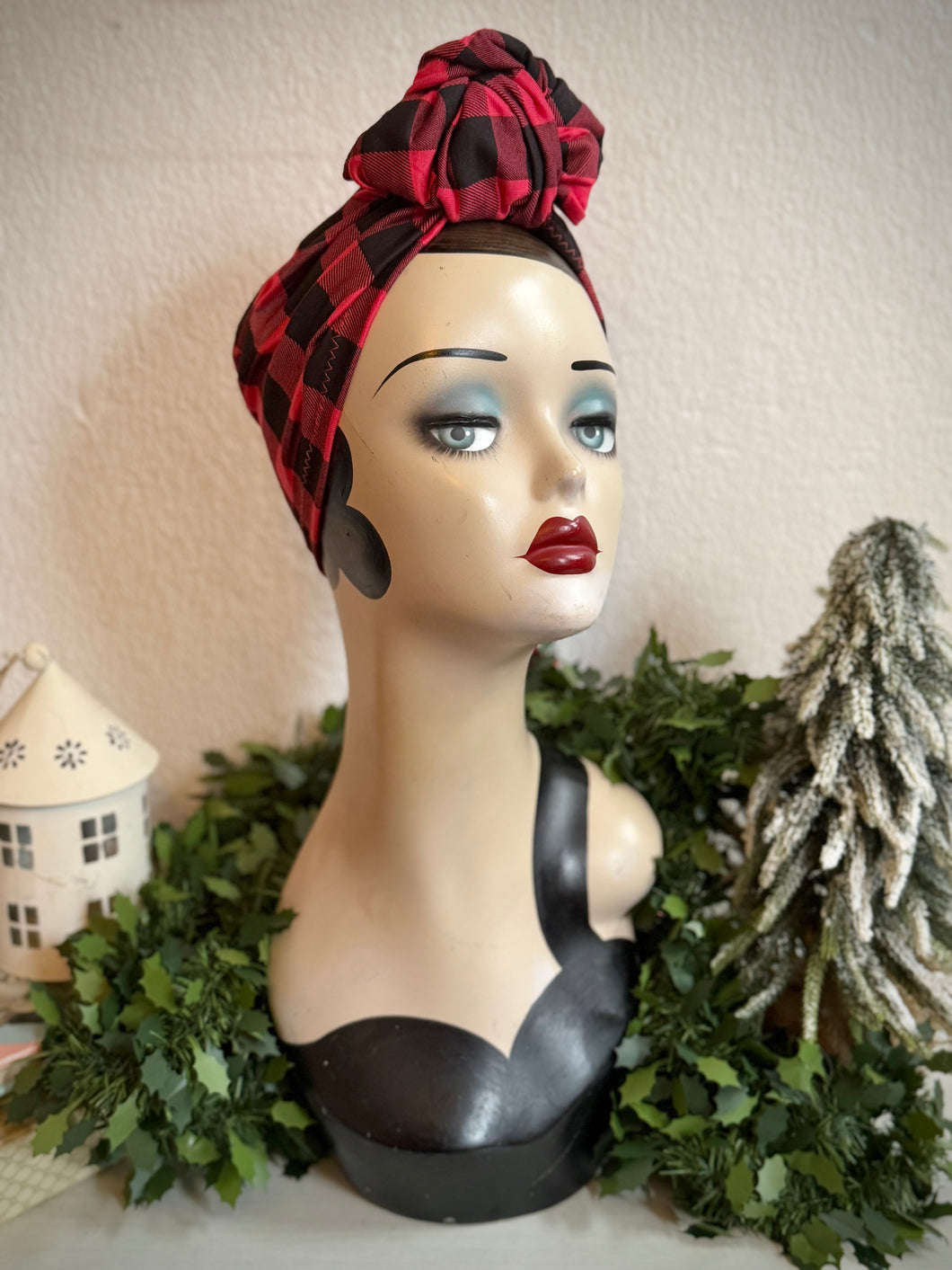 Mannequin head with a red and black plaid headscarf against a neutral background with Christmas decorative elements.