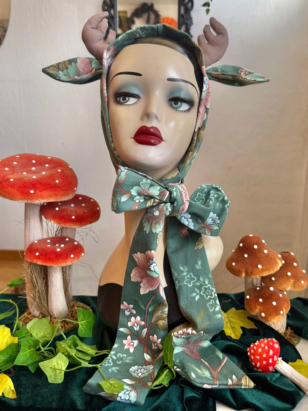 Mannequin head with floral deer antlers surrounded by decorative mushrooms and leaves.