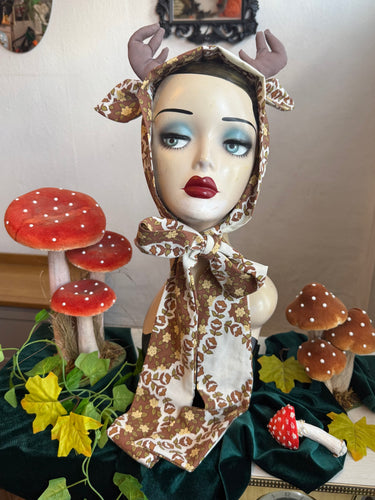 Mannequin head with deer antlers and a patterned scarf, surrounded by decorative mushrooms and leaves.